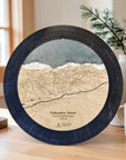 A round wooden cribbage board game featuring an engraved map of Palisades Tahoe, displayed on a wooden table in a cozy room setting with a window, plant, and mug in the background.