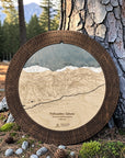 Round wooden cribbage board featuring an engraved, laser-cut ski map of Palisades Tahoe, displayed outdoors leaning against a large tree trunk in a forest setting.
