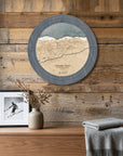 A circular wooden Palisades Tahoe ski map cribbage board that doubles as a wall art piece. styled on a rustic wood-paneled wall above a wooden console table with a framed photo, a vase with dried branches, and a folded gray blanket.