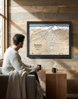 Man sitting on a couch in a cozy ski cabin with a framed map of Revelstoke Mountain on the wall.