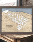 Framed 3d wood map of Ski Santa Fe in New Mexico on a wooden surface inside a rustic ski cabin.