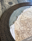 Close-up of a circular wooden cribbage board game with layered ski trail map details and a dark wood segmented playing track, photographed on a rustic wood surface.