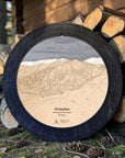 Matte Onyx Whitefish ski map cribbage board staged beside stacked firewood in a mountain cabin setting.