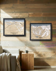 Two framed ski trail maps on a wooden wall above a sofa in a cozy ski cabin.