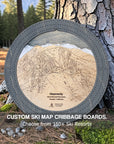 Handcrafted Ski Trail Map Cribbage Board with a dark grey 3-player cribbage track, featuring an engraved wooden map of a Ski Resort, displayed leaning against a tree in a forest setting.