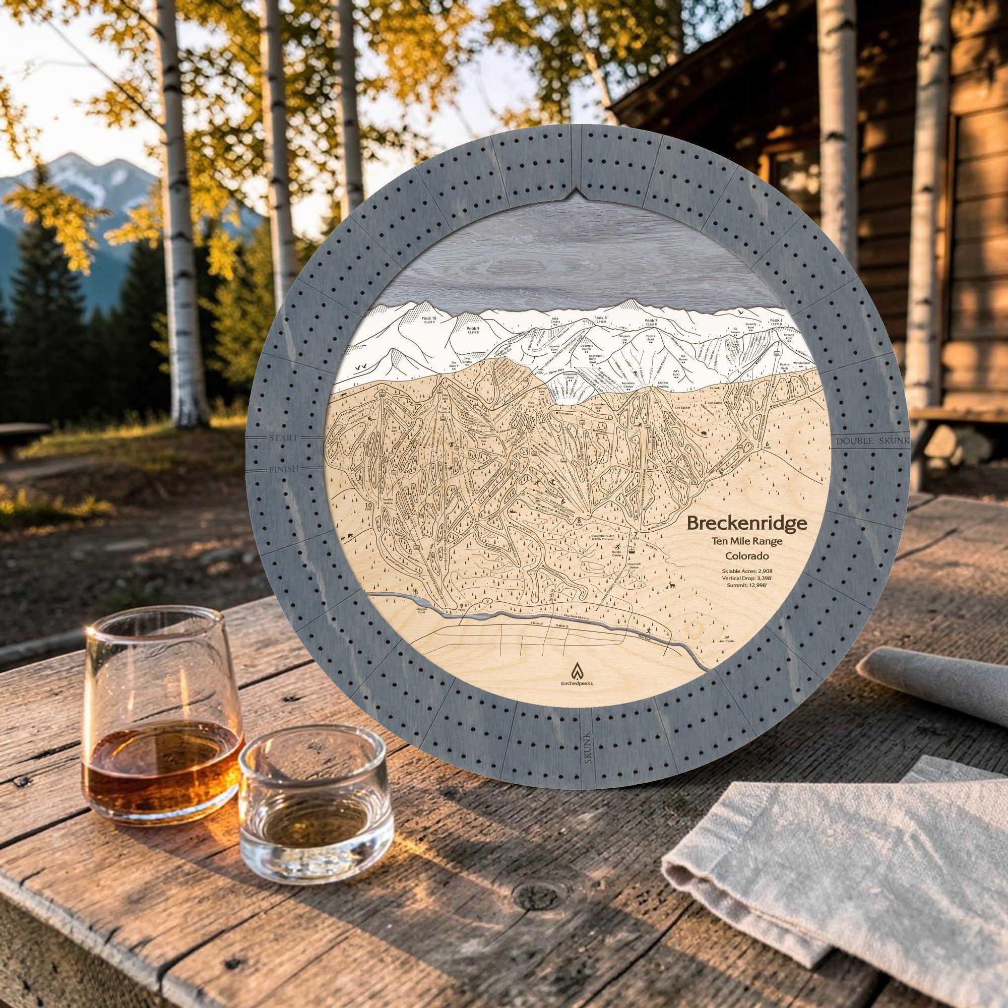 Graphite Grey Breckenridge ski map cribbage board on an outdoor cabin table beside whiskey glasses.