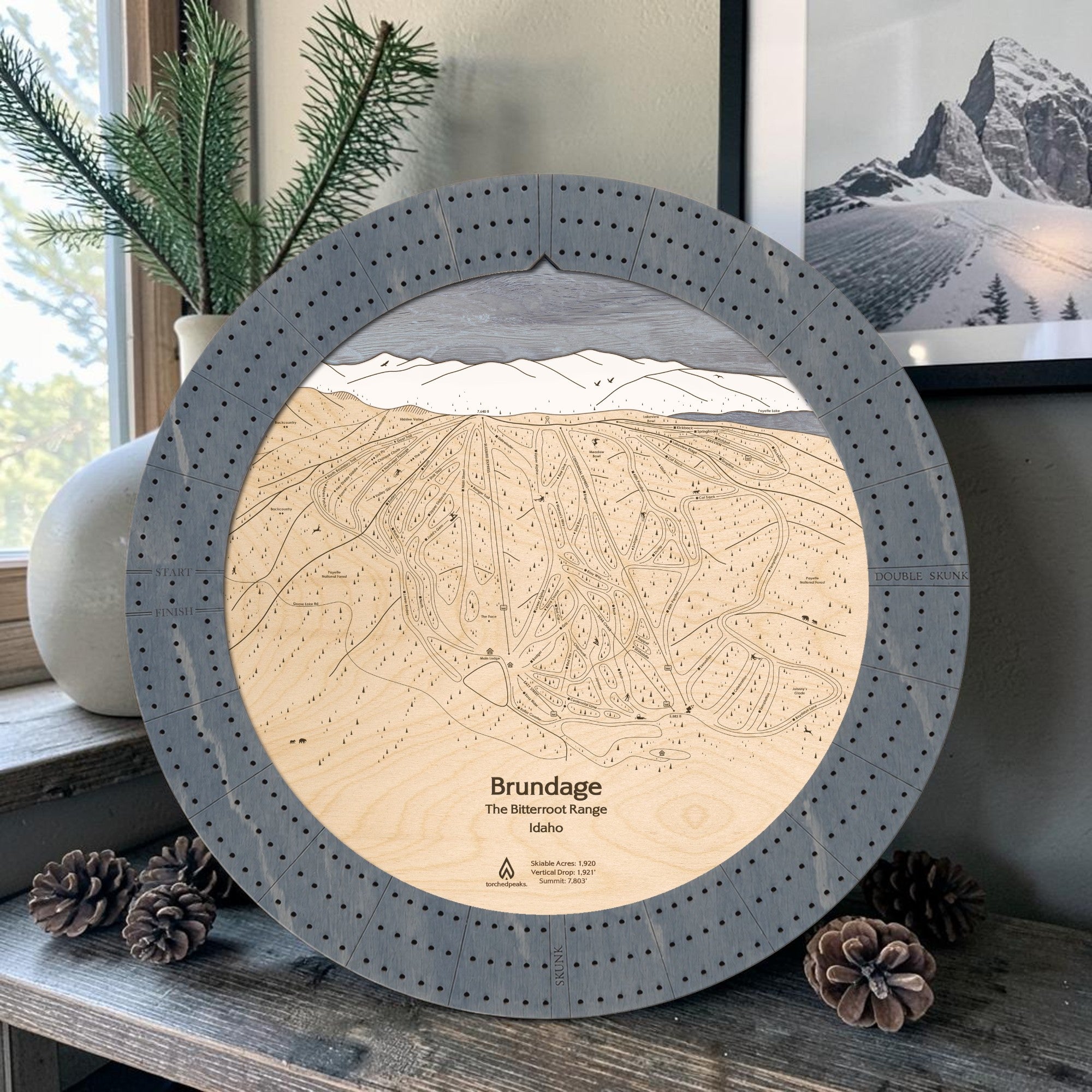 Graphite Grey Brundage Mountain wooden cribbage board displayed beside pinecones and a framed mountain photograph.