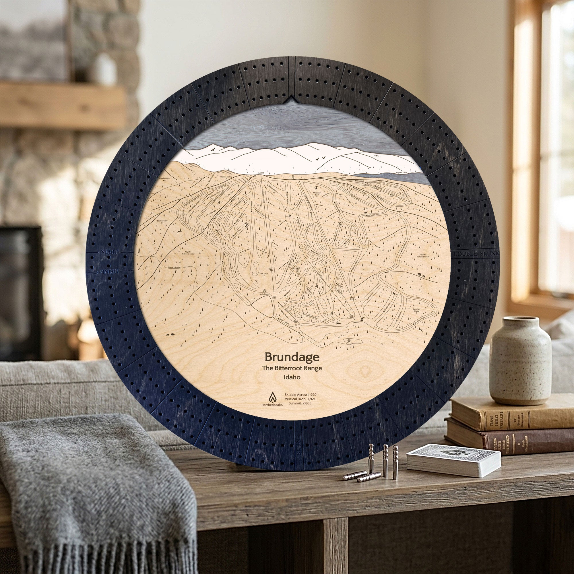Matte Onyx Brundage Mountain ski map cribbage board displayed beside books and a gray blanket in a bright cabin interior.