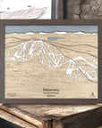 Wooden trail map of Discovery Ski Area on a rustic wooden surface inside a cozy ski cabin. 