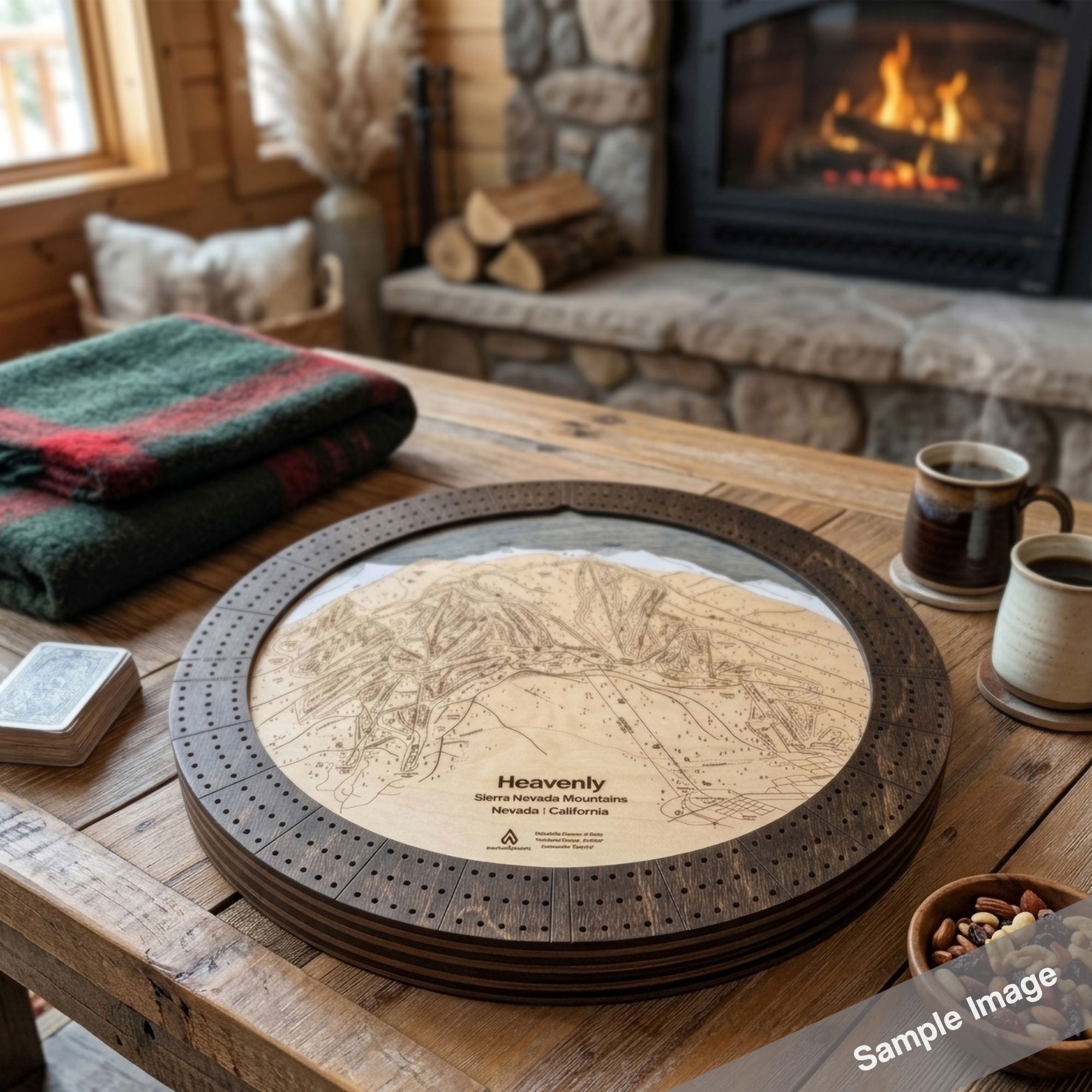 A round wooden cribbage board game set displayed on a rustic wooden table in a cozy living room setting with a stone fireplace and a burning fire in the background. The cribbage board includes a dark brown circular outer frame with pre-drilled  cribbage peg holes and a light birch inner board featuring a detailed ski map design.
