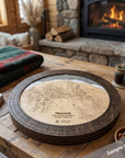 A round wooden cribbage board game set displayed on a rustic wooden table in a cozy living room setting with a stone fireplace and a burning fire in the background. The cribbage board includes a dark brown circular outer frame with pre-drilled  cribbage peg holes and a light birch inner board featuring a detailed ski map design.