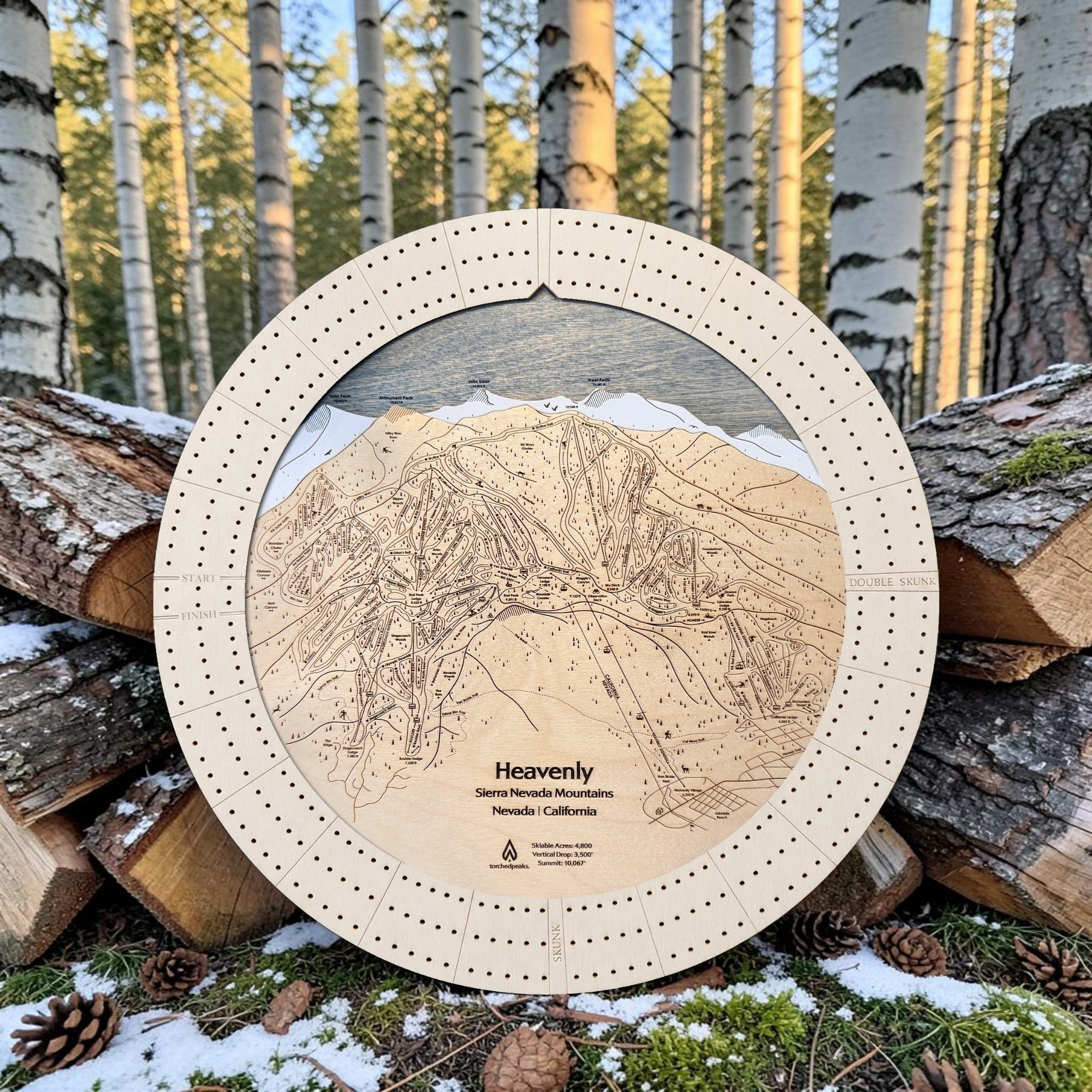 Handcrafted Heavenly Ski Trail Map Cribbage Board with a natural maple cribbage track, featuring an engraved wooden map of Heavenly Ski Resort, displayed outdoors on stacked logs in a snowy alpine forest.