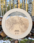Handcrafted Heavenly Ski Trail Map Cribbage Board with a natural maple cribbage track, featuring an engraved wooden map of Heavenly Ski Resort, displayed outdoors on stacked logs in a snowy alpine forest.