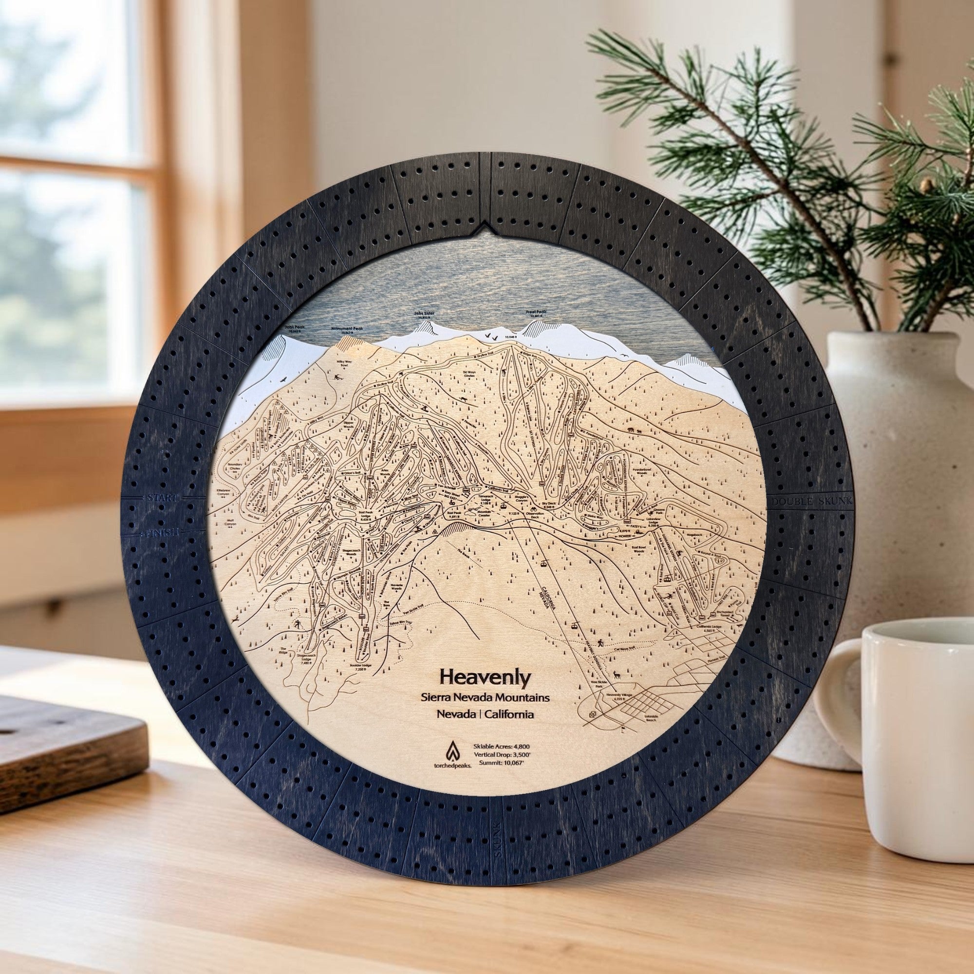 Handcrafted Heavenly Ski Trail Map Cribbage Board with a black walnut cribbage track, featuring an engraved wooden map of Heavenly Ski Resort, displayed on a mountain-inspired tabletop beside a coffee mug and evergreen arrangement.