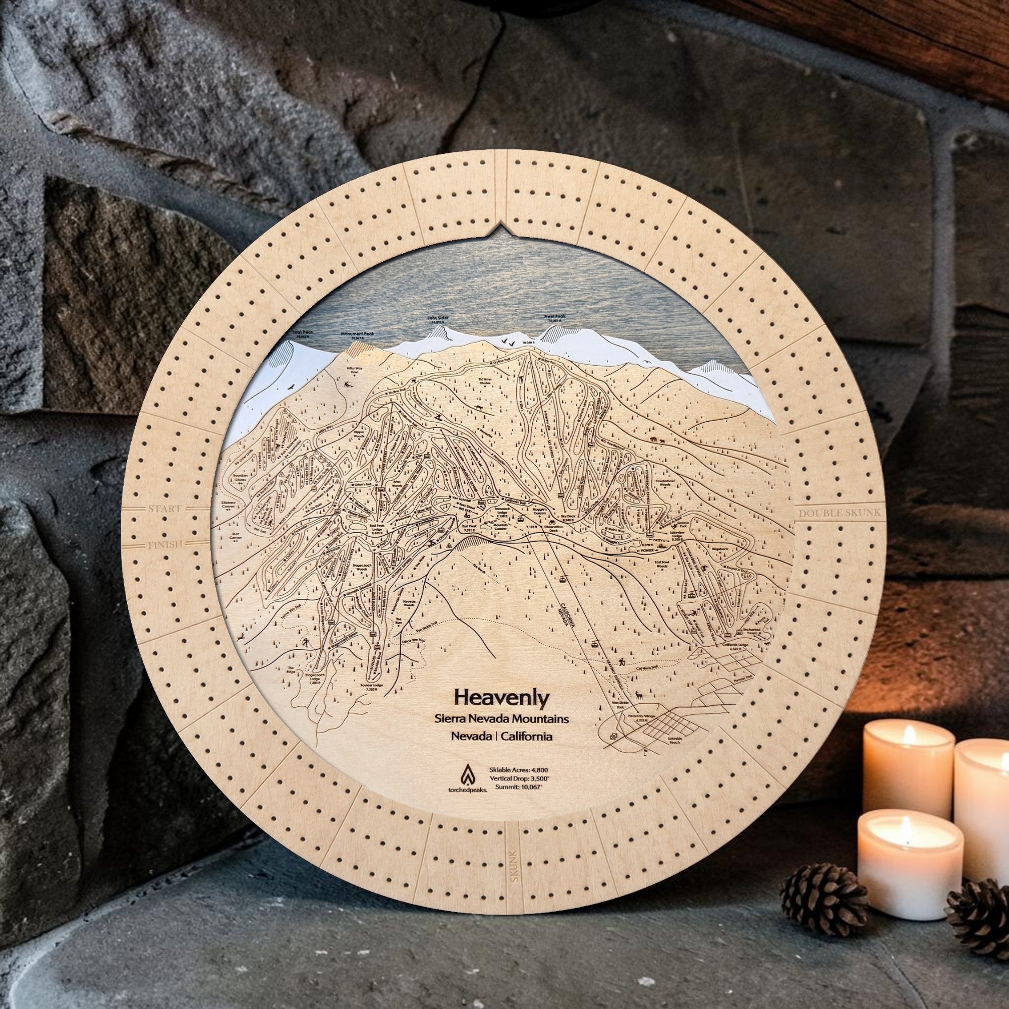 Handcrafted Heavenly Ski Trail Map Cribbage Board with a black walnut cribbage track, featuring an engraved wooden map of Heavenly Ski Resort, displayed on a mountain cabin tabletop with evergreen branches and a coffee mug.