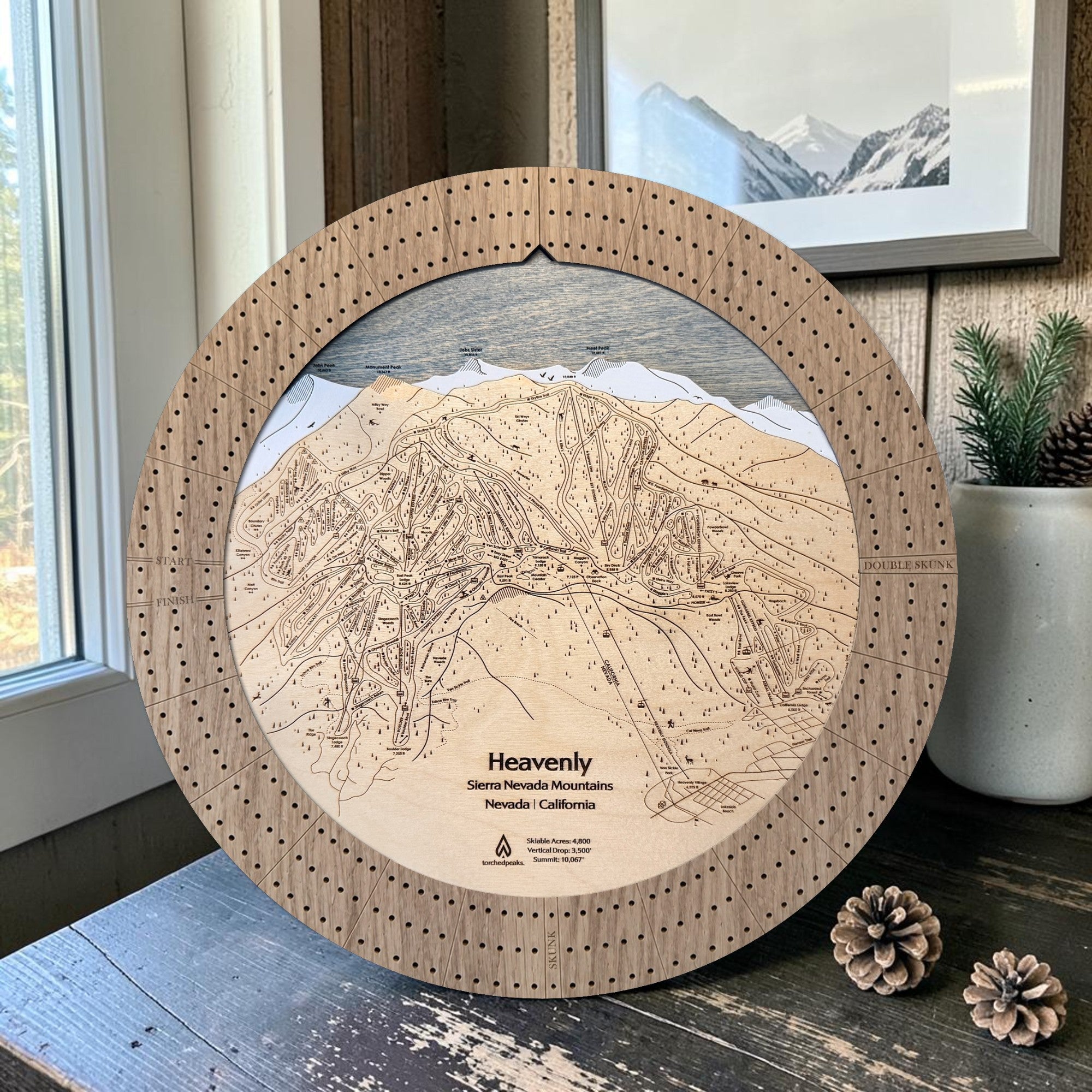 Handcrafted Heavenly Ski Trail Map Cribbage Board with a natural oak cribbage track, featuring an engraved wooden map of Heavenly Ski Resort, displayed on a mountain-inspired tabletop with pinecones, evergreen décor, and framed alpine artwork.