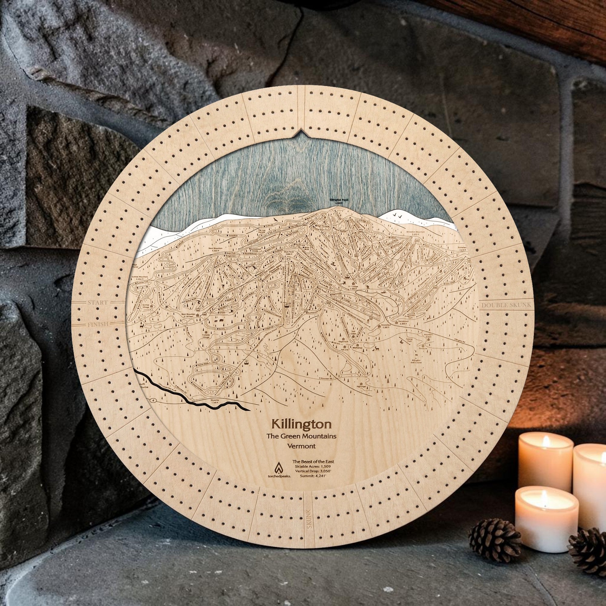 Killington wooden ski map cribbage board in Birch beside a stone fireplace with candles and pinecones.