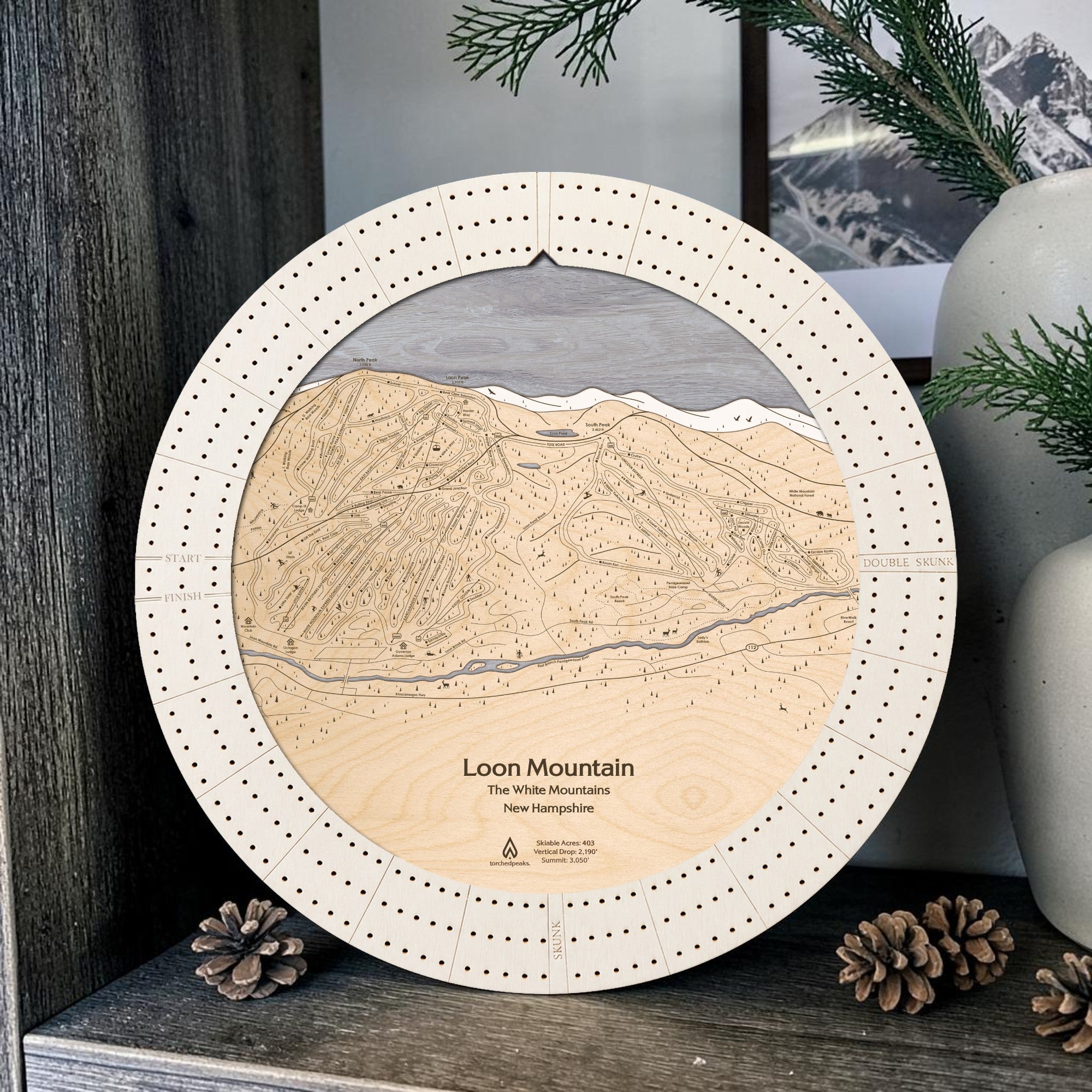 Arctic White Loon Mountain ski map cribbage board displayed with pinecones and greenery on a rustic shelf in a ski cabin.