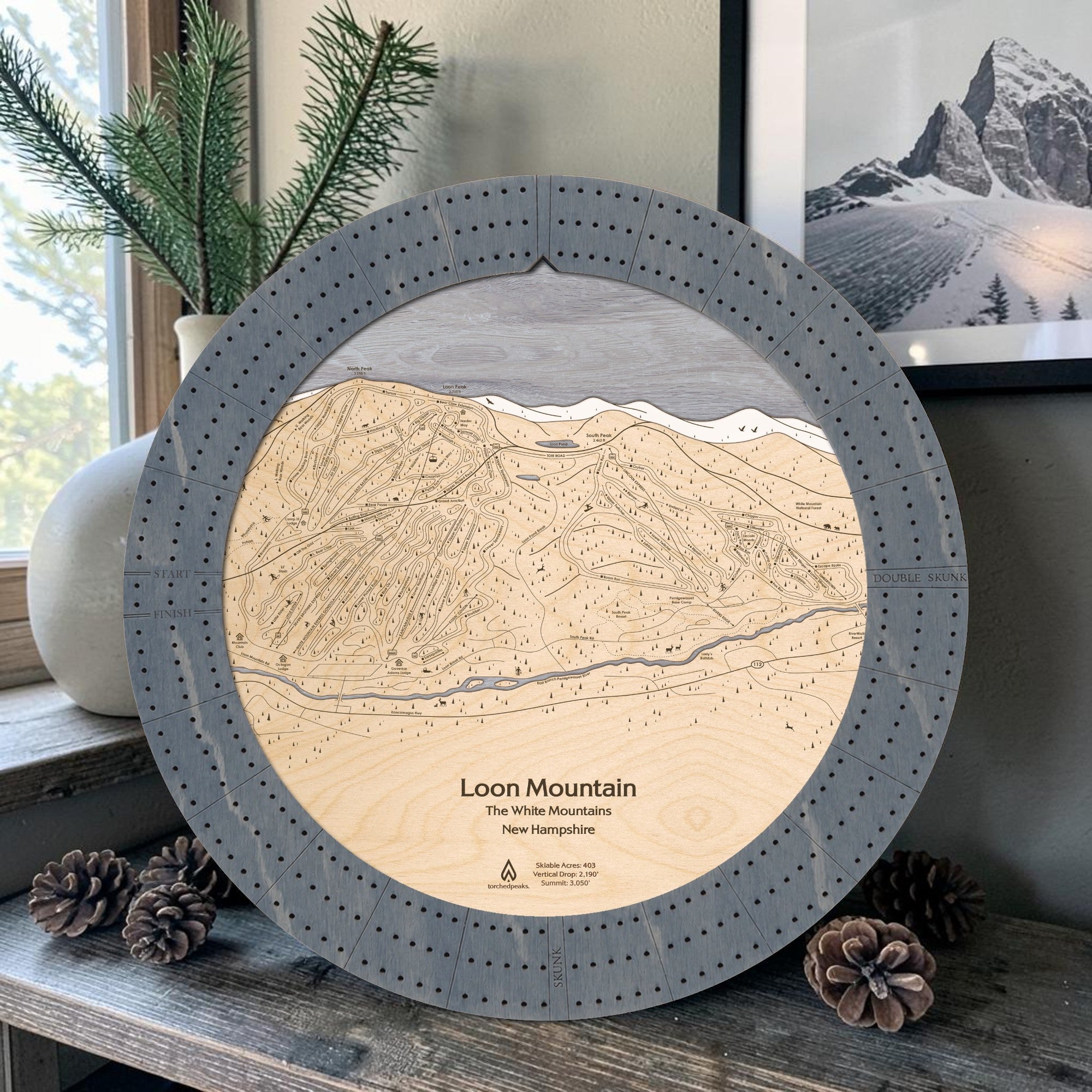 Graphite Grey Loon Mountain wooden cribbage board displayed with pinecones and a framed mountain photograph in a modern ski condo. 