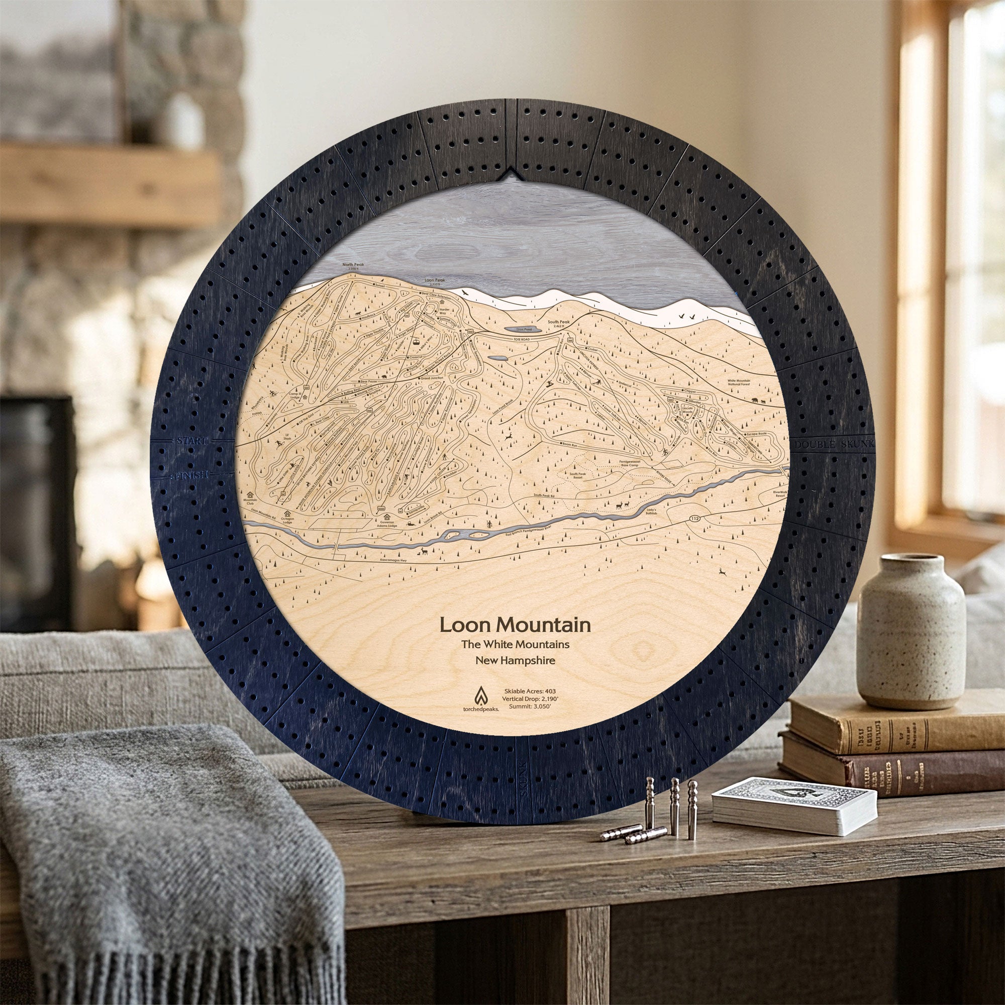 Matte Onyx Loon Mountain ski map cribbage board displayed beside books and a gray blanket as ski cabin décor.