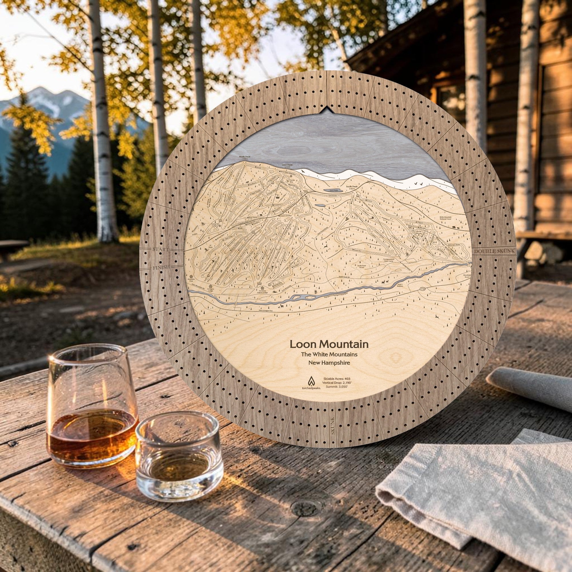 Weathered Timber Loon Mountain ski map cribbage board displayed on an outdoor cabin table beside whiskey glasses.