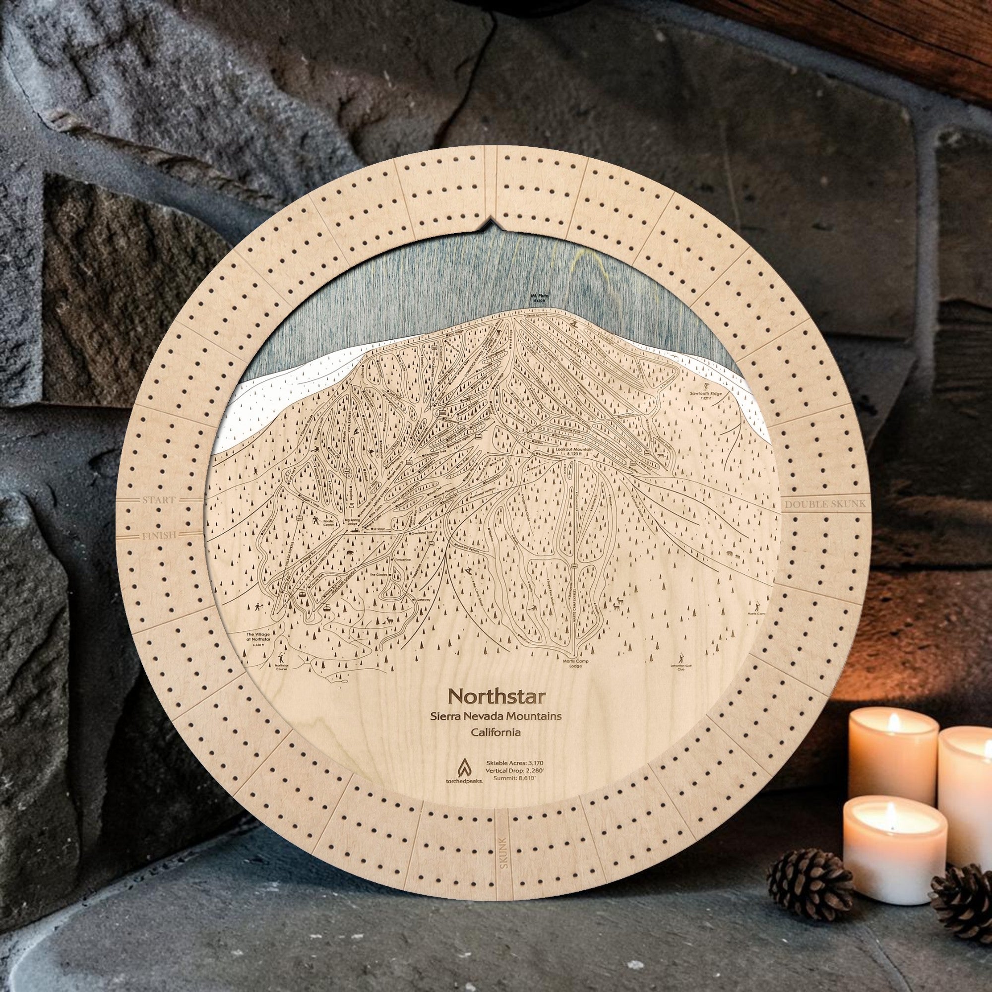 Northstar wood cribbage board in Birch beside a stone fireplace with candles.