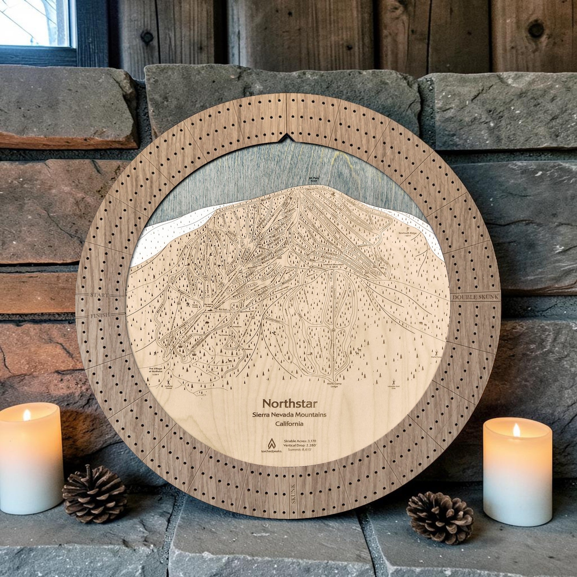 Northstar cribbage board in Weathered Timber beside a stone fireplace with candles and pinecones.