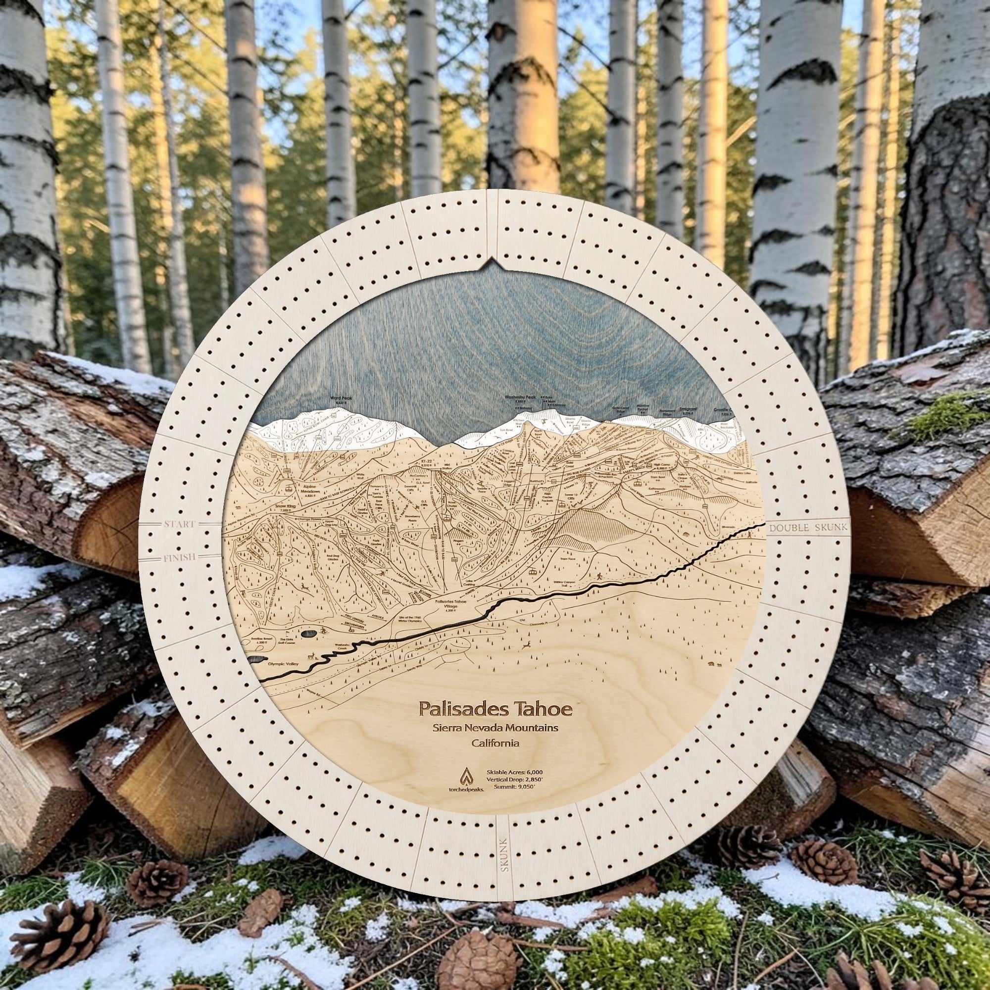 Round wooden ski map cribbage of Palisades Tahoe, Sierra Nevada Mountains, displayed outdoors on stacked logs with birch trees and snow in the background.