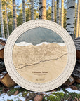 Round wooden ski map cribbage of Palisades Tahoe, Sierra Nevada Mountains, displayed outdoors on stacked logs with birch trees and snow in the background.