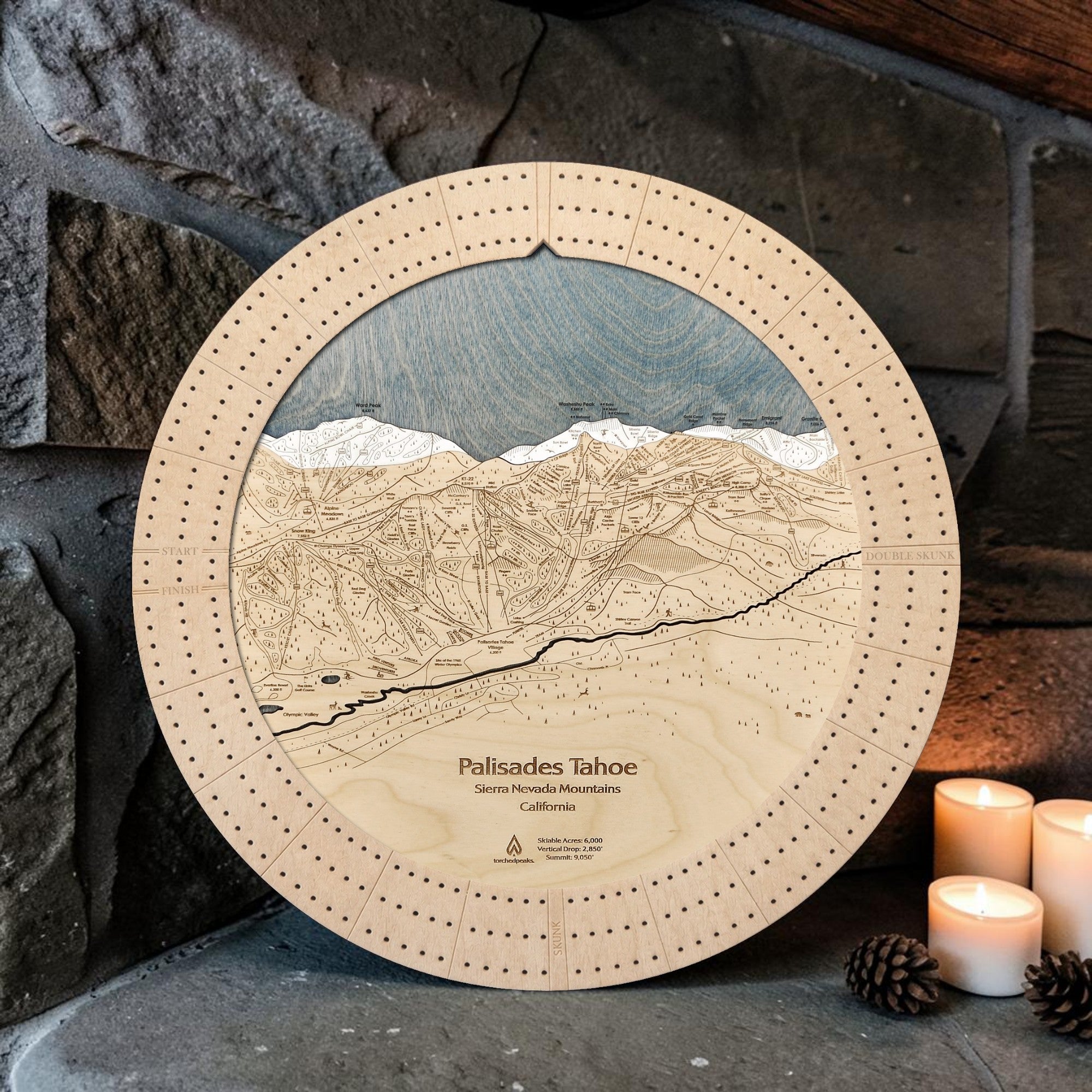 A 16" round wooden cribbage  featuring an engraved detailed ski map of Palisades Tahoe ski resort, set against a rustic stone wall with lit candles and pinecones.