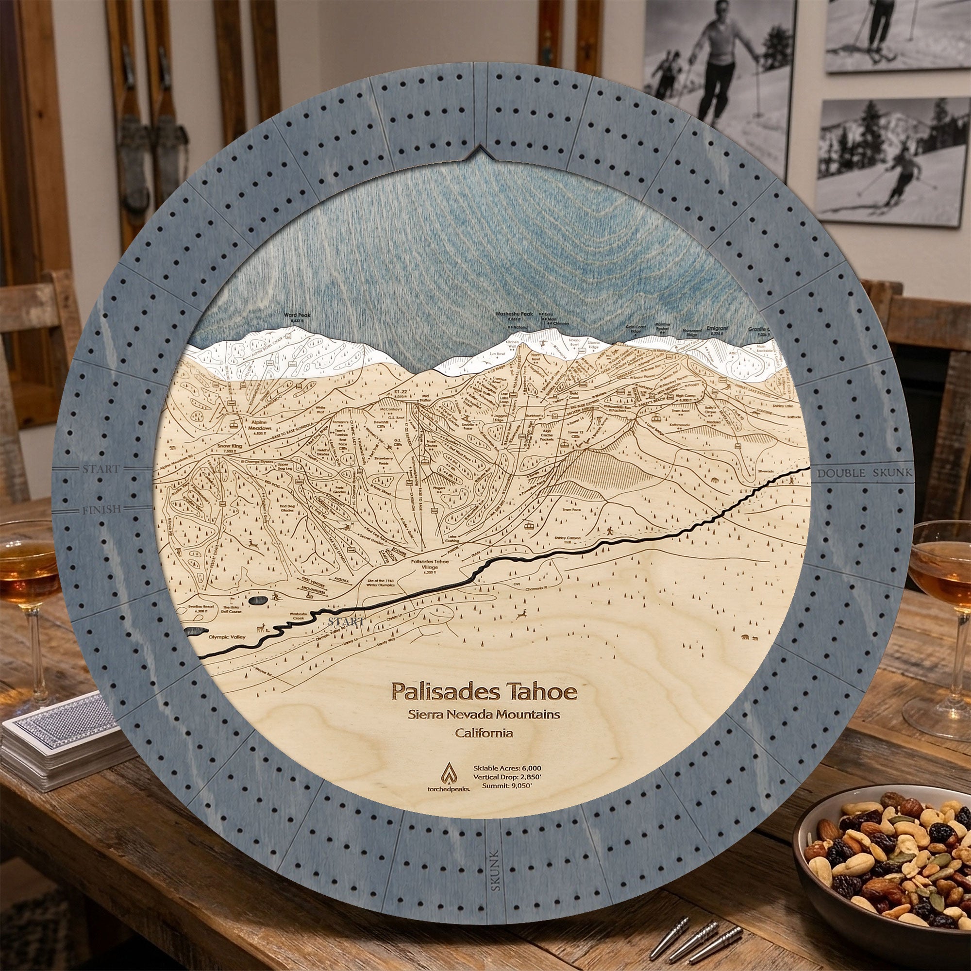 A 16" round wooden Palisades Tahoe map cribbage board game displayed on a rustic wooden table. The plaque features a detailed 3D wood-cut terrain design with a blue-gray outer ring, beige wood layers for the mountain slopes, and a blue-gray sky layer at the top.