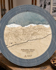 A 16" round wooden Palisades Tahoe map cribbage board game displayed on a rustic wooden table. The plaque features a detailed 3D wood-cut terrain design with a blue-gray outer ring, beige wood layers for the mountain slopes, and a blue-gray sky layer at the top.