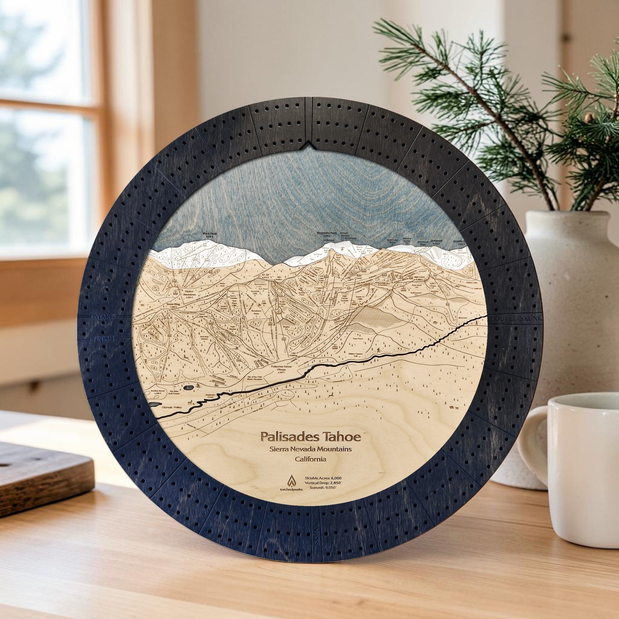A round wooden cribbage board game featuring an engraved map of Palisades Tahoe, displayed on a wooden table in a cozy room setting with a window, plant, and mug in the background.