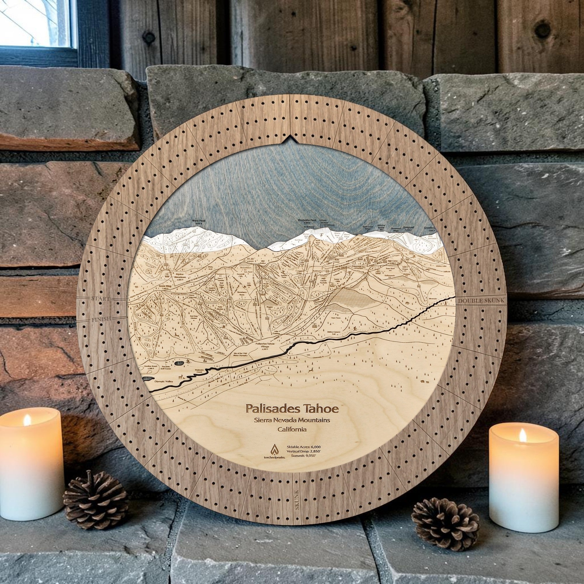 Round wooden ski trail map cribbage board featuring an engraved topographic mountain map of Palisades Tahoe ski resort. styled on a stone shelf with two lit candles and pinecones.