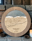 Round wooden ski trail map cribbage board featuring an engraved topographic mountain map of Palisades Tahoe ski resort. styled on a stone shelf with two lit candles and pinecones.