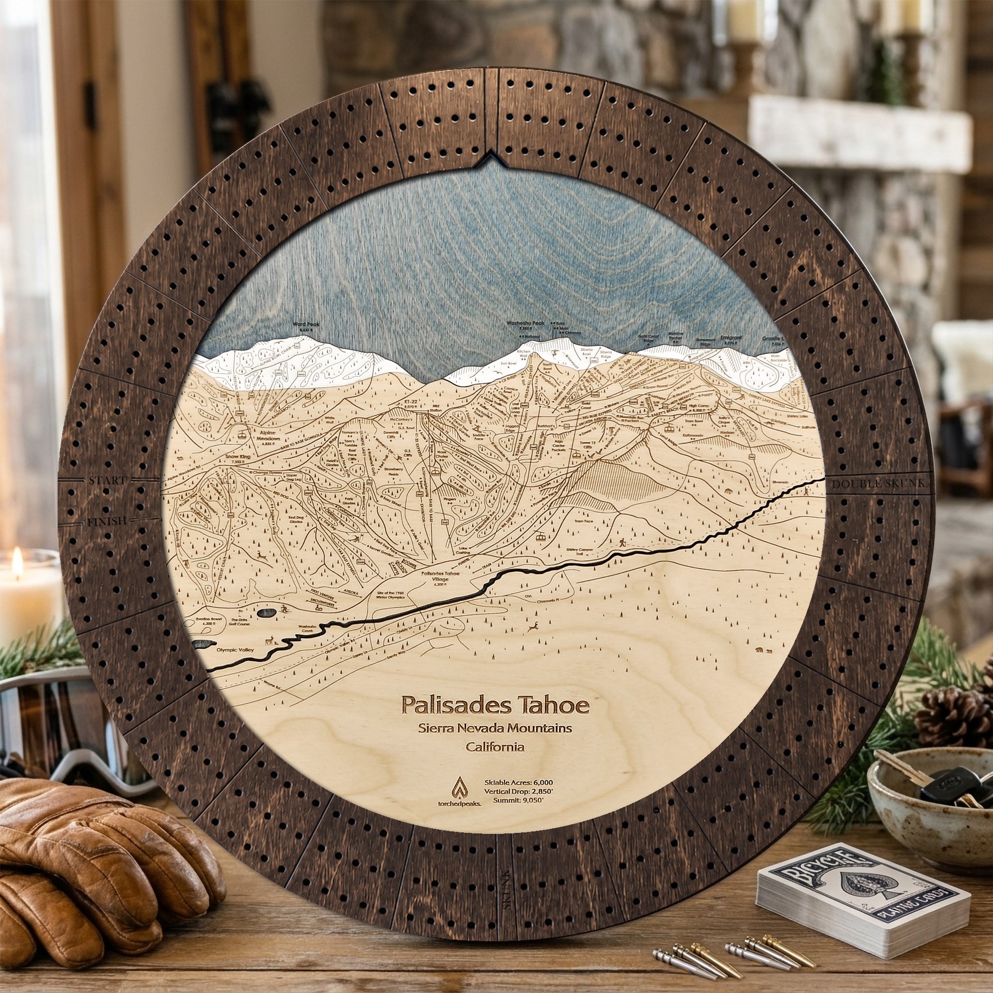 A round wooden Palisades Tahoe cribbage board game displayed on a rustic wooden table in a cozy room setting. The board features a detailed ski map of the mountain slopes in the center, surrounded by a dark brown perforated outer ring.