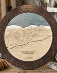 A round wooden Palisades Tahoe cribbage board game displayed on a rustic wooden table in a cozy room setting. The board features a detailed ski map of the mountain slopes in the center, surrounded by a dark brown perforated outer ring.