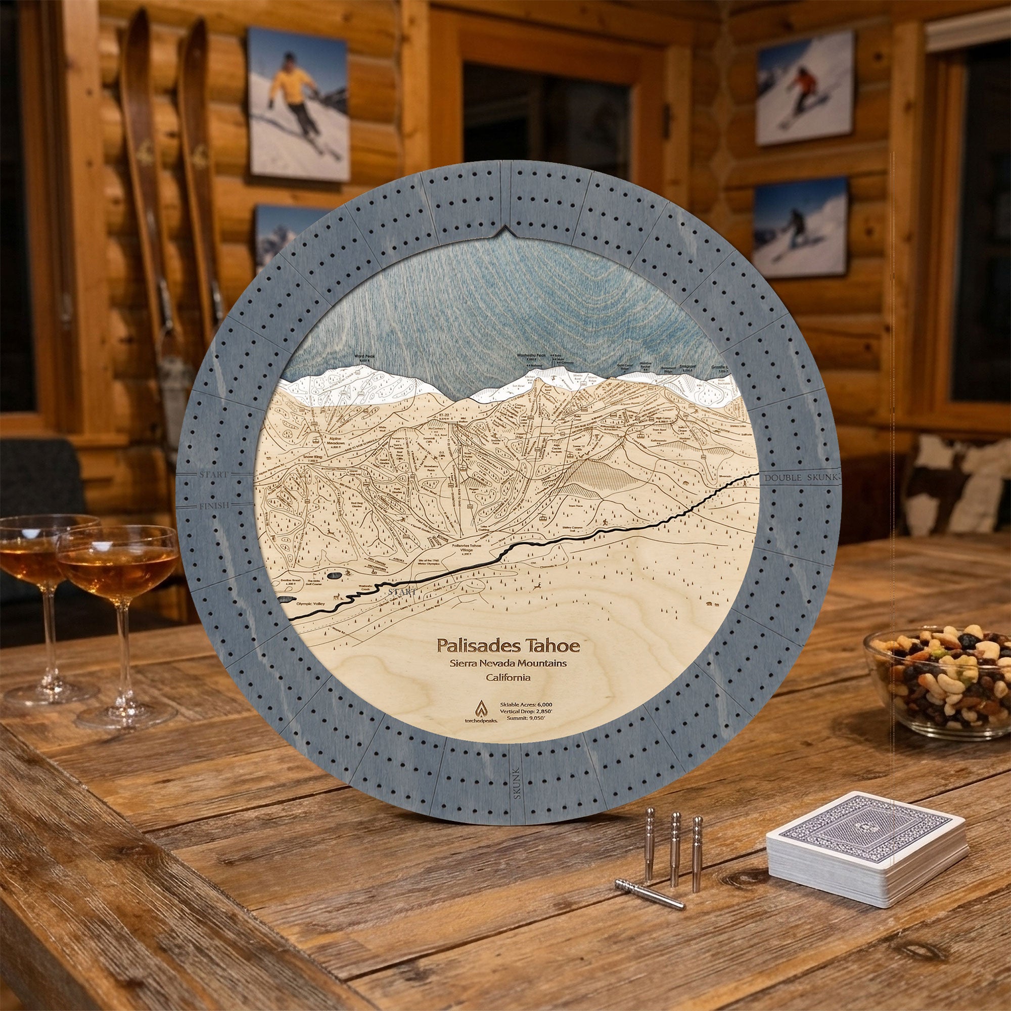 A circular wooden cribbage board game titled 'Pallisades Tahoe' displayed on a rustic wooden table in a cozy lodge setting. The board features a detailed, engraved map of the ski resort, with a graphite grey outer ring, white and blue snow, and beige wood-toned terrain.