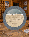A circular wooden cribbage board game titled 'Pallisades Tahoe' displayed on a rustic wooden table in a cozy lodge setting. The board features a detailed, engraved map of the ski resort, with a graphite grey outer ring, white and blue snow, and beige wood-toned terrain.
