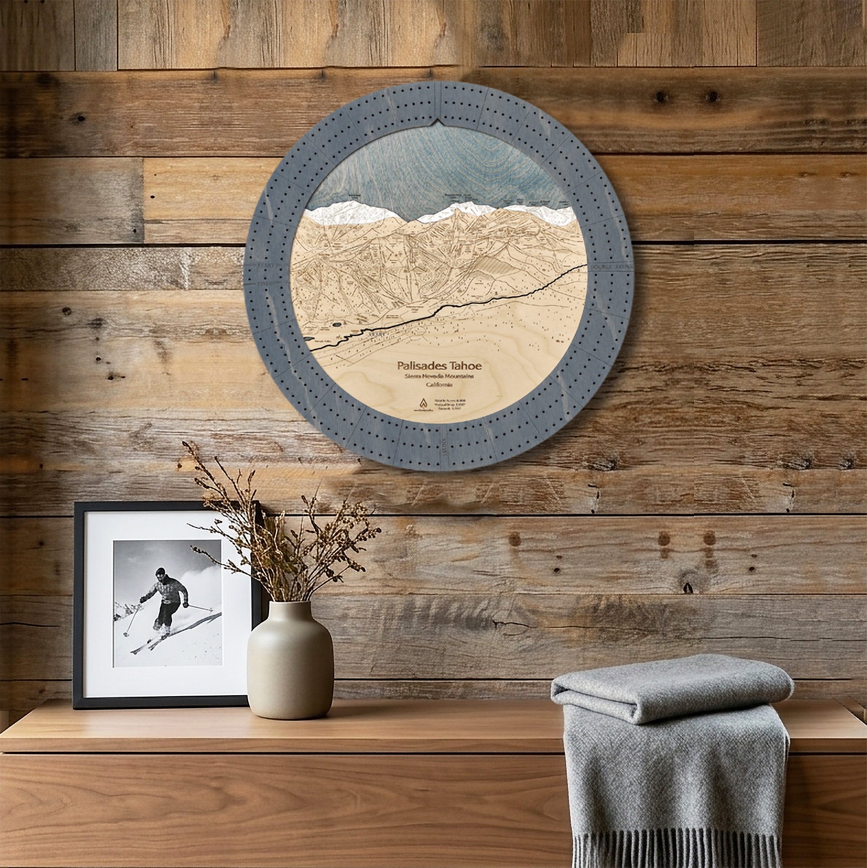 A circular wooden Palisades Tahoe ski map cribbage board that doubles as a wall art piece.  styled on a rustic wood-paneled wall above a wooden console table with a framed photo, a vase with dried branches, and a folded gray blanket.