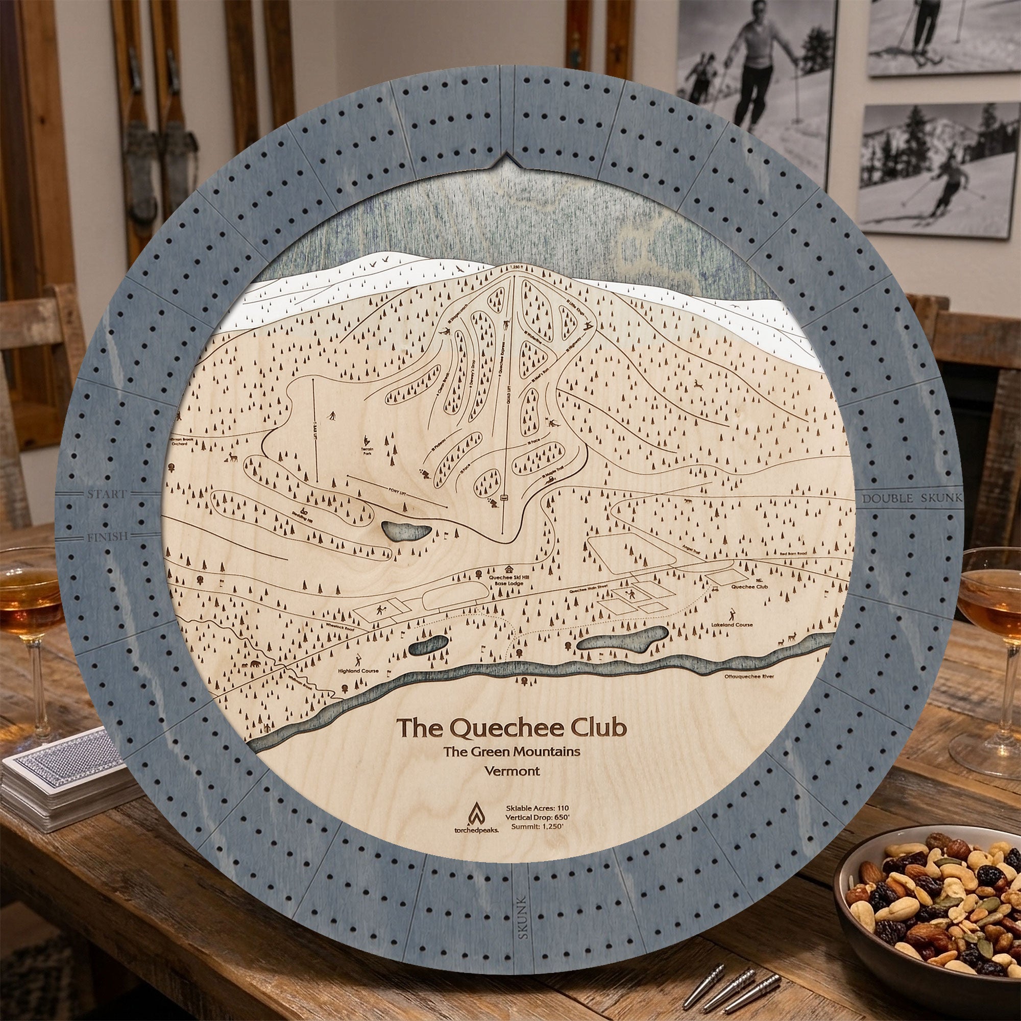 Graphite Grey Quechee Club wooden ski map cribbage board staged on a table inside a rustic ski cabin.