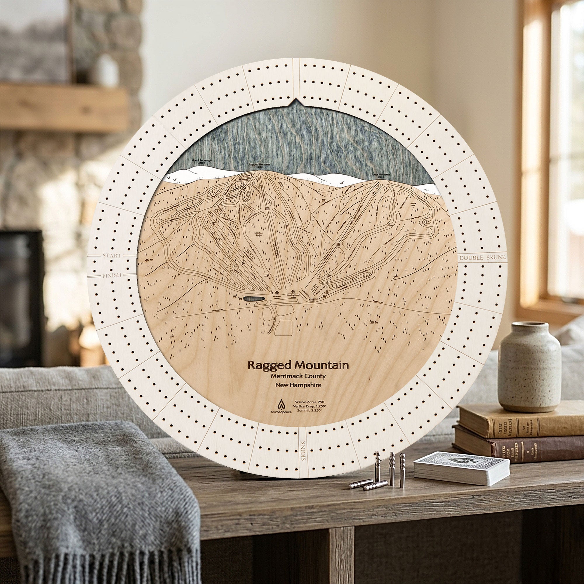 A round wooden map cribbage board of Ragged Mountain ski area, New Hampshire, styled on a rustic wooden table with a gray throw blanket, books, and a small vase in a cozy indoor setting.
