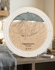 A round wooden map cribbage board of Ragged Mountain ski area, New Hampshire, styled on a rustic wooden table with a gray throw blanket, books, and a small vase in a cozy indoor setting.