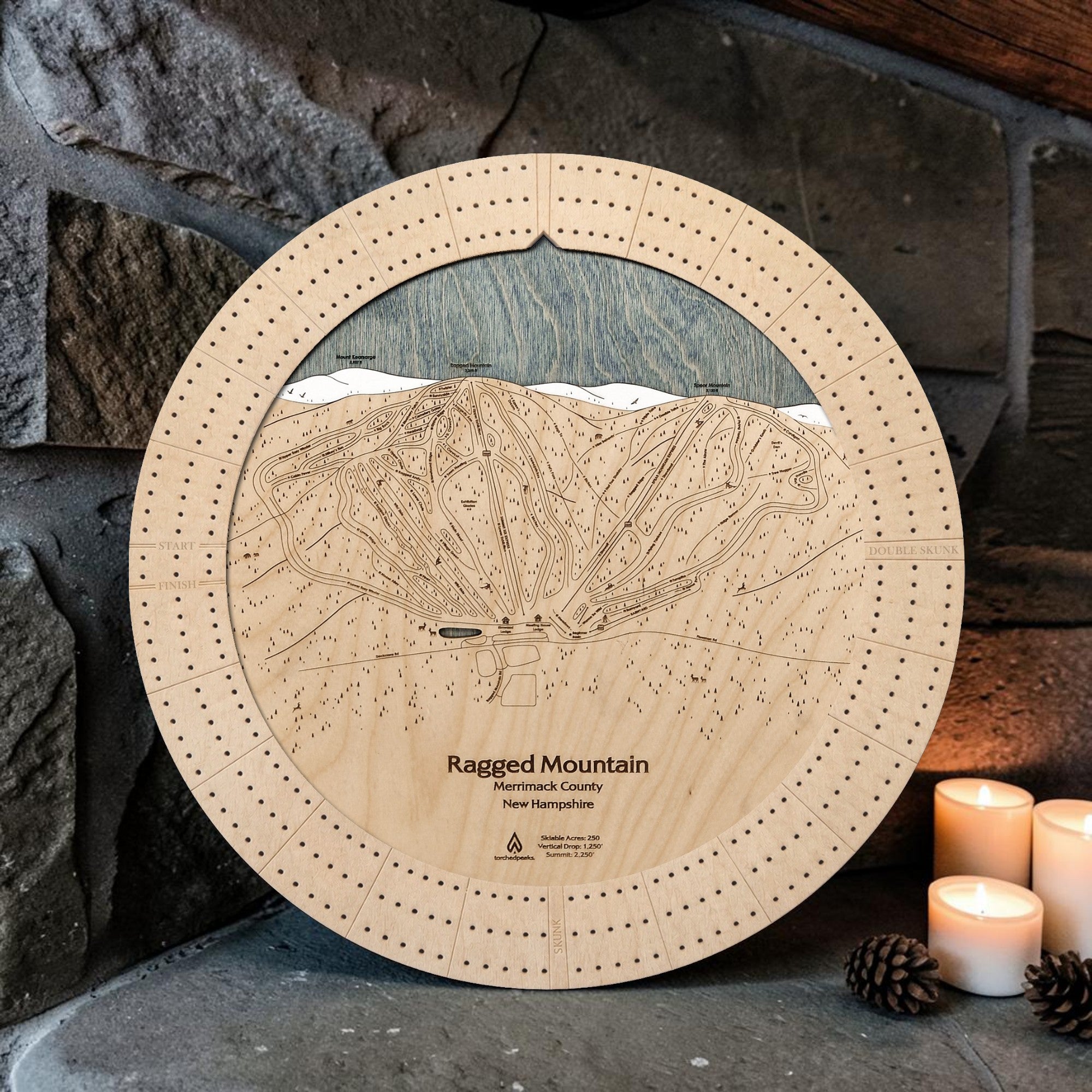 Ragged Mountain wooden ski map cribbage board in Birch beside a stone fireplace with candles and pinecones.
