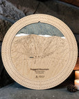 Ragged Mountain wooden ski map cribbage board in Birch beside a stone fireplace with candles and pinecones.