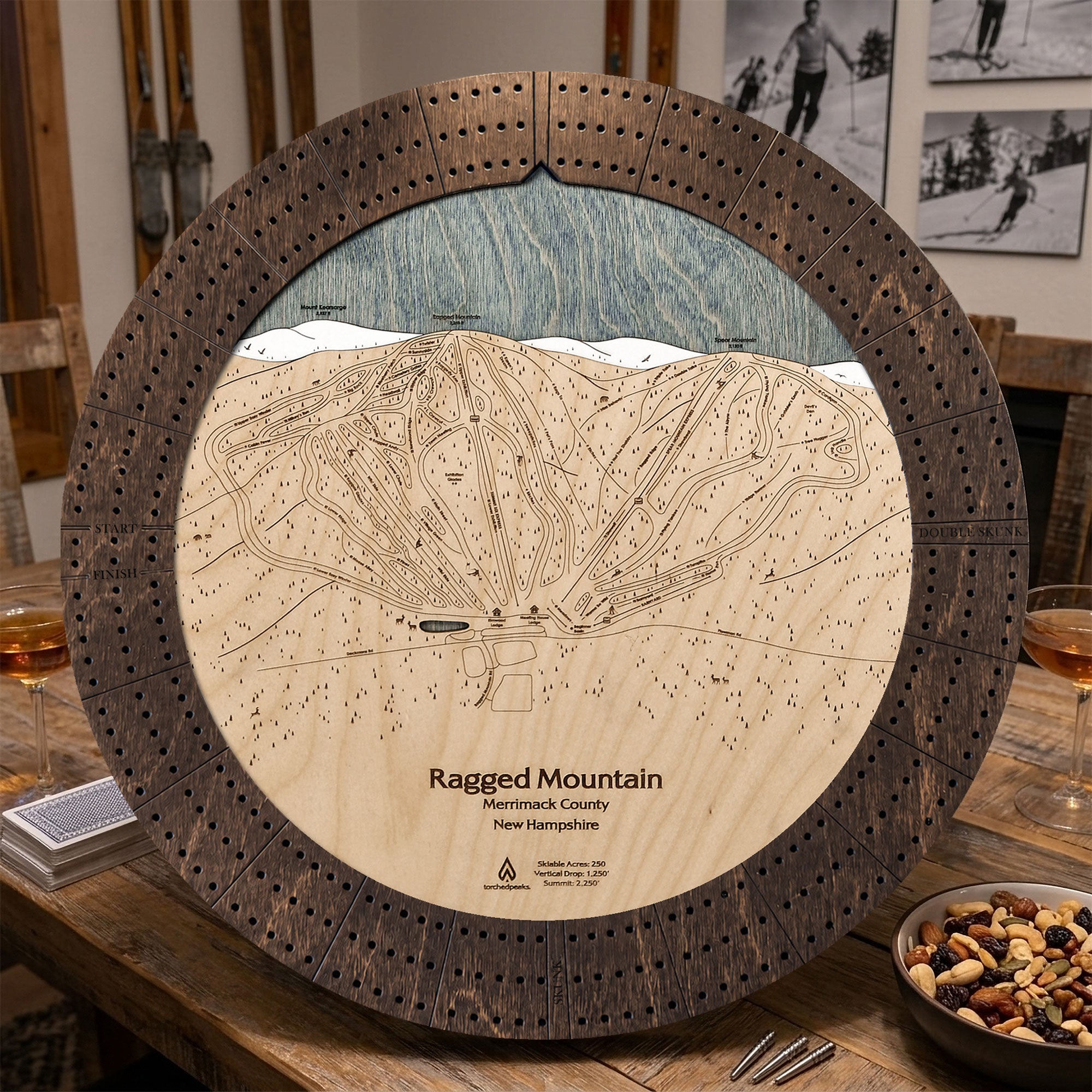 Ragged Mountain ski map cribbage board in Rustic Mocha styled on a distressed table with playing cards inside rustic ski cabin. 