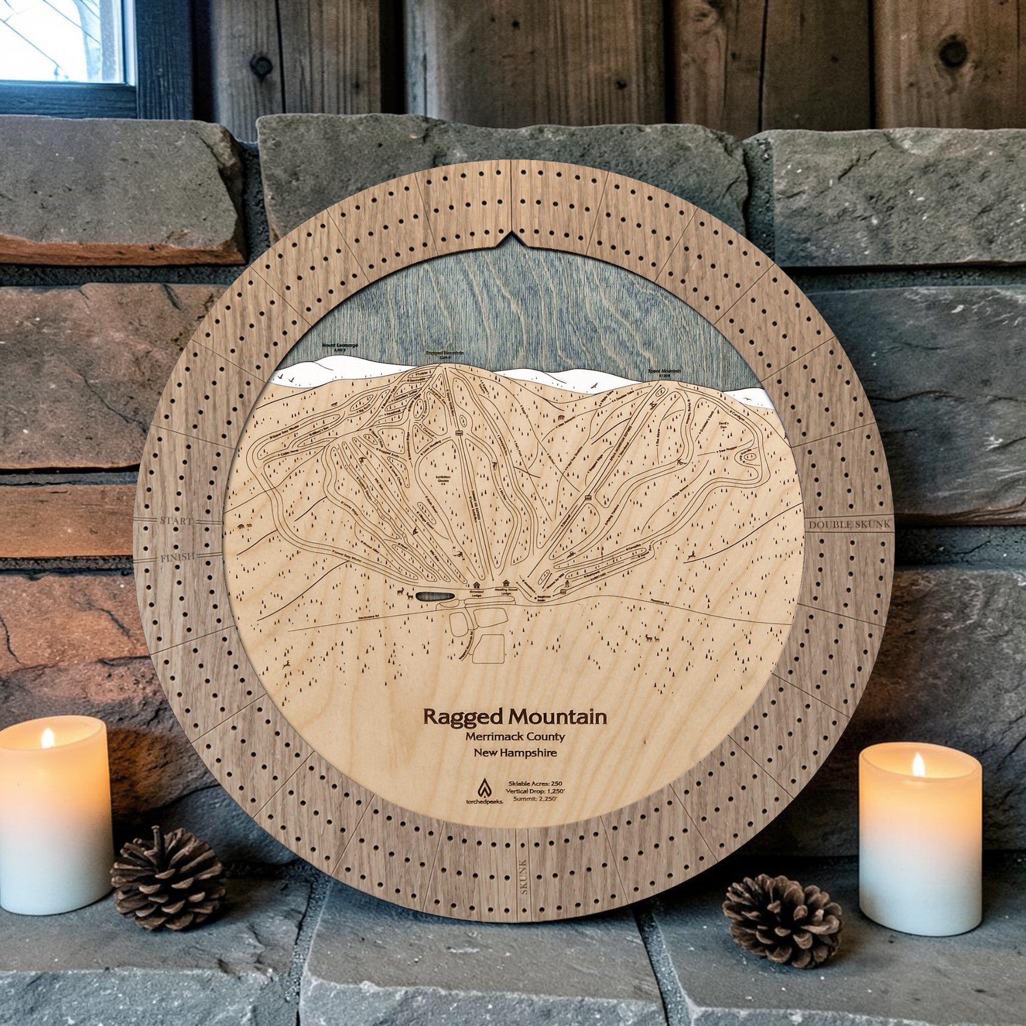 Ragged Mountain cribbage board in Weathered Timber beside a stone fireplace with candles and pinecones.