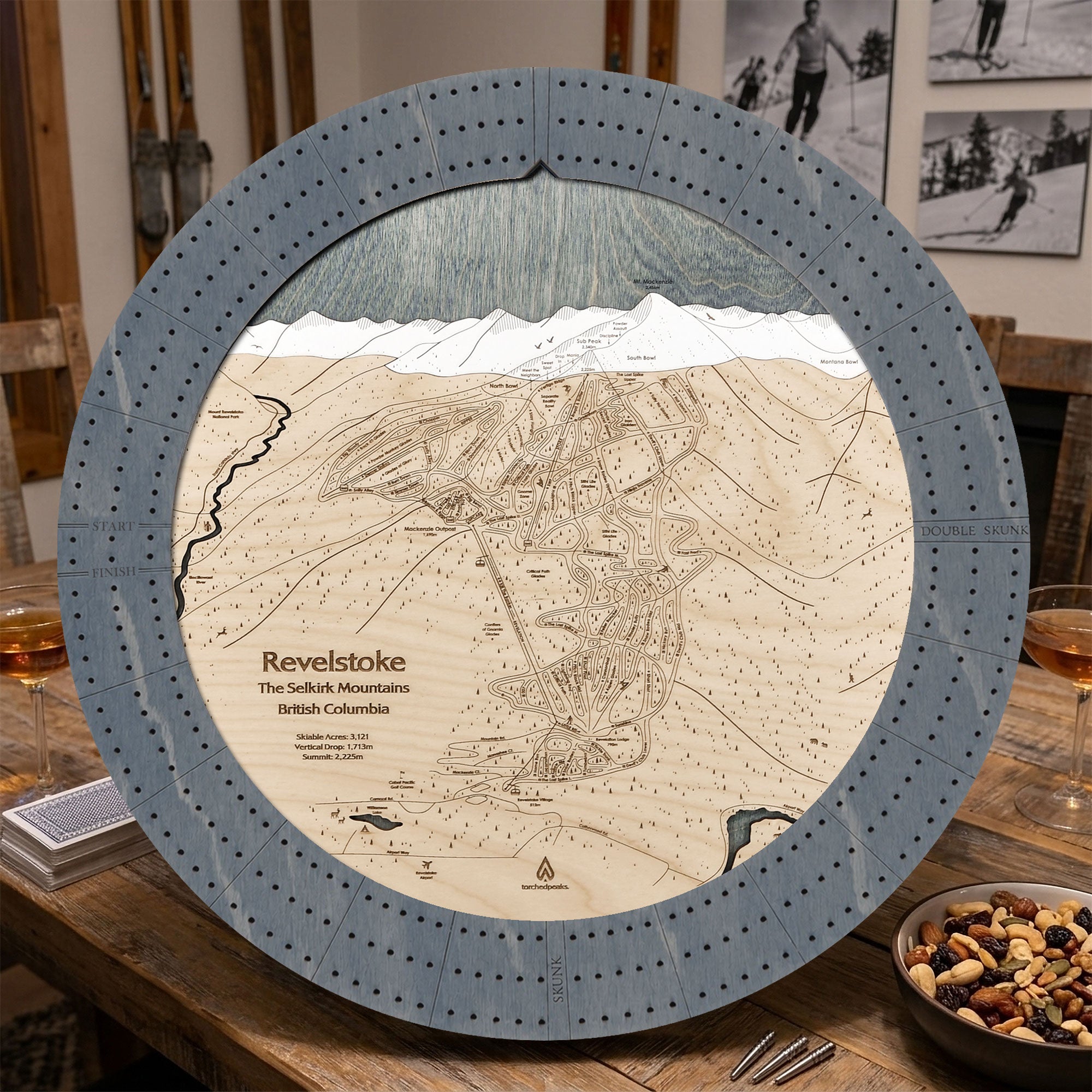 Graphite Grey Revelstoke ski map cribbage board staged on a wooden table inside a rustic mountain cabin.
