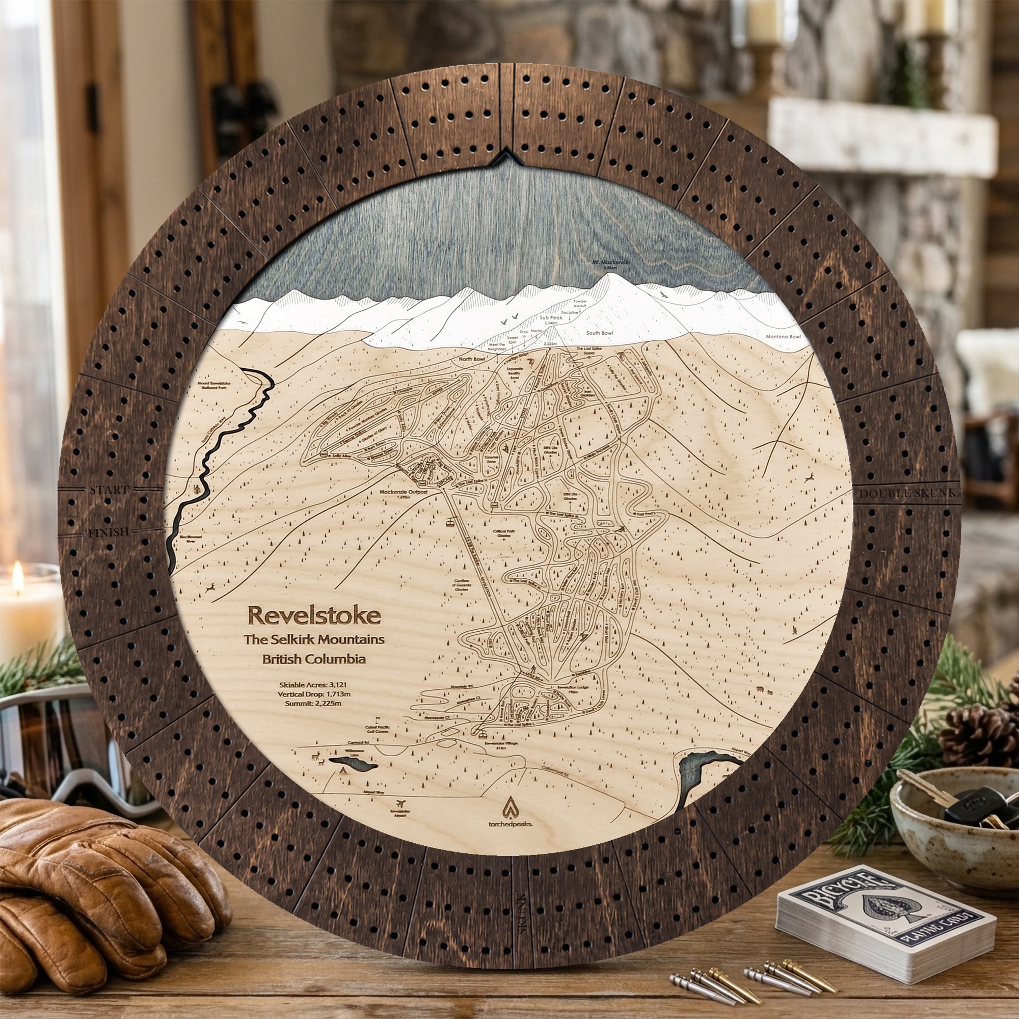 Rustic Mocha Revelstoke ski map cribbage board displayed with playing cards on a table in a cozy mountain lodge.