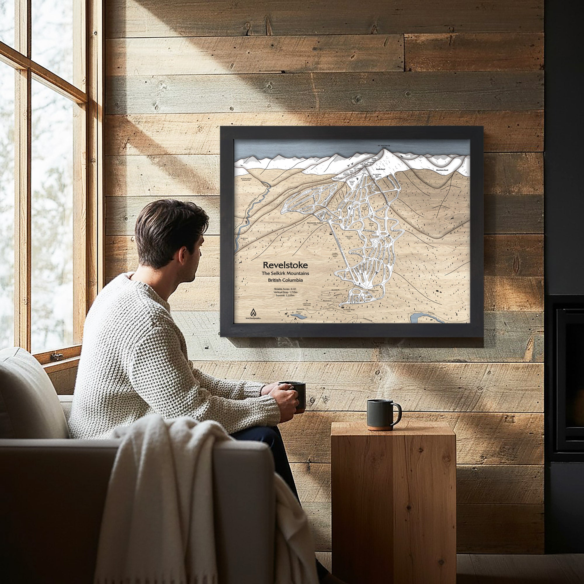 Man sitting on a couch in a cozy ski cabin with a framed map of Revelstoke Mountain on the wall.